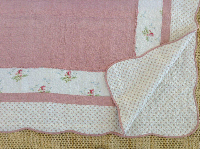A close-up of a quilted textile, likely a baby blanket or quilt, featuring a dusty pink solid section, white borders with small floral prints, and a cream-colored dotted reverse side with pink piping.