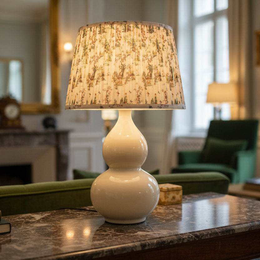 White lamp with a patterned shade on a marble table in a room with green furniture and a fireplace.