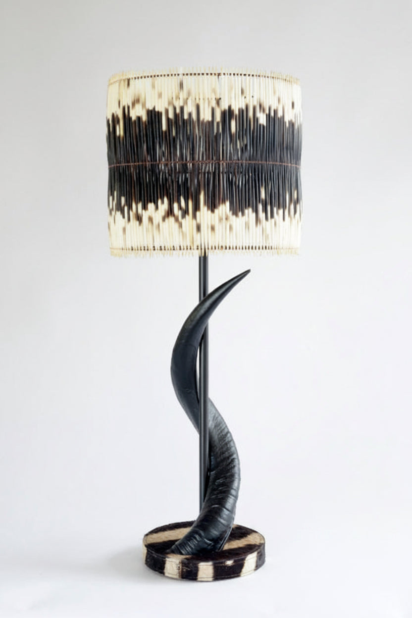 A sculptural table lamp with a black horn-like curved base, a black metal stem, and a drum shade made of vertical black and cream animal-print material, set against a plain white background.
